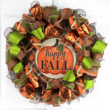 Round brown orange and green mesh wreath with a circle metal sign in the middle with a pumpkin and the words Happy Fall, hanging on a white door